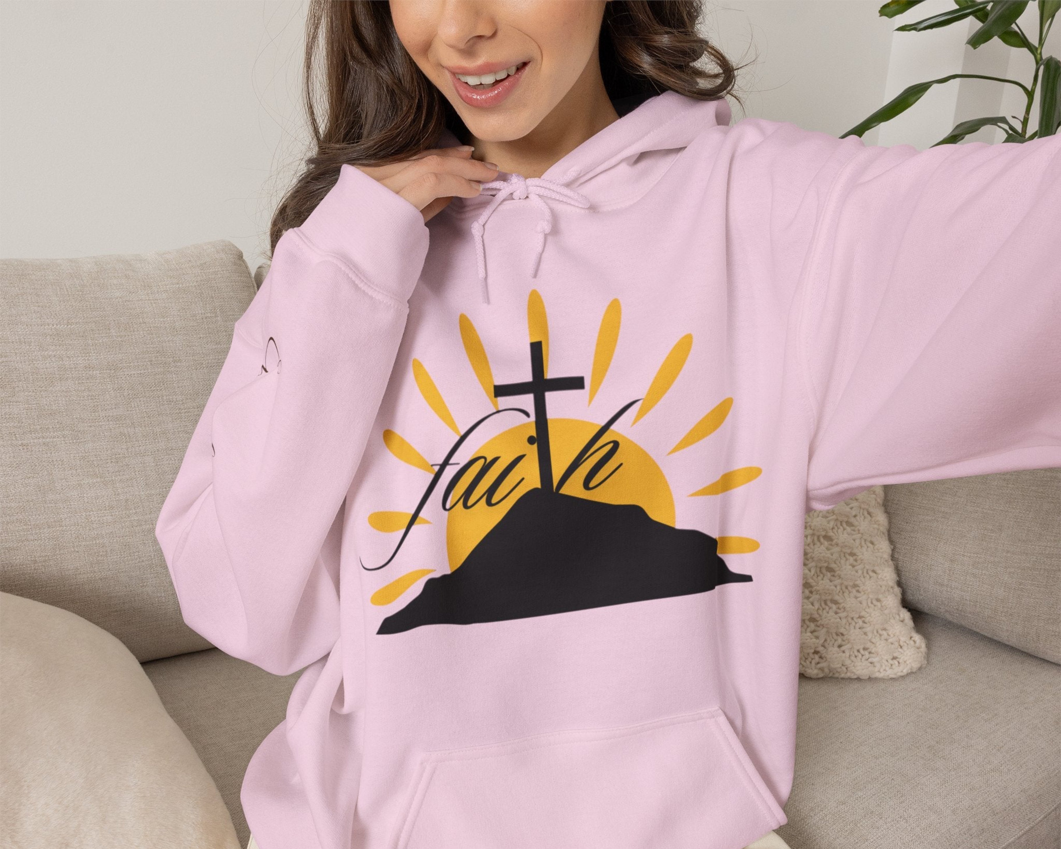 Person wearing a pink hoodie with a graphic design of a cross, sun, and the word 'faith' sitting on a couch.